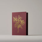 Christmas card featuring a gold-outlined Hellebore  on a burgundy background, standing on a neutral surface.