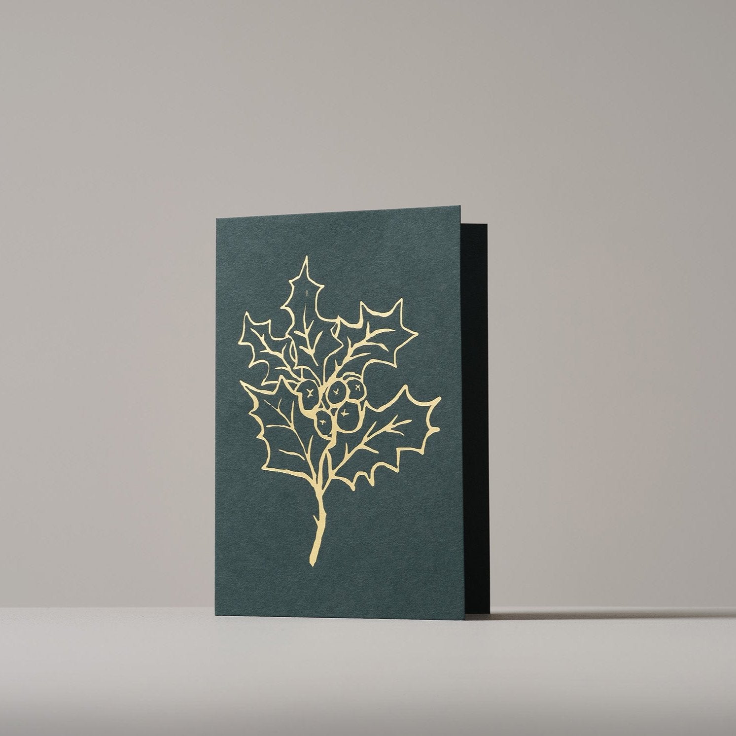 Christmas card featuring a gold-outlined holly branch with berries on a dark green background, standing on a neutral surface