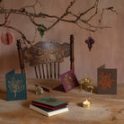 
A rustic holiday scene with hand-made Christmas cards and paper ornaments. An ornate wooden chair sits against a neutral, earthy-toned wall, with a branch hanging above adorned with pastel paper ornaments in muted green, pink, and purple hues. On the wooden table in front, three cards are displayed upright, featuring simple botanical illustrations in gold and orange ink, one with a holly leaf, another with mistletoe, and the third with a floral design. A small stack of blank cards lies beside a bottle of i